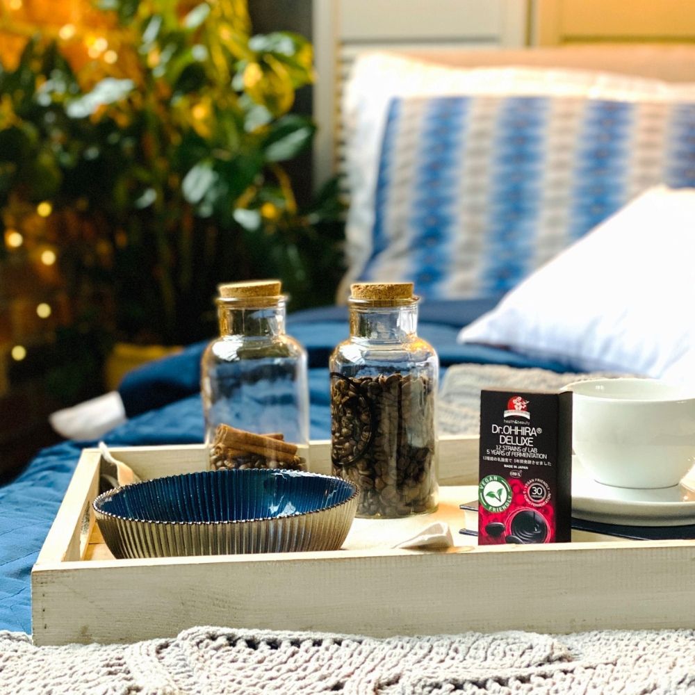 Cutie Dr.Ohhira Deluxe pe tavă lemn, alături de bol albastru și borcane cu cafea, în dormitor decorat elegant.