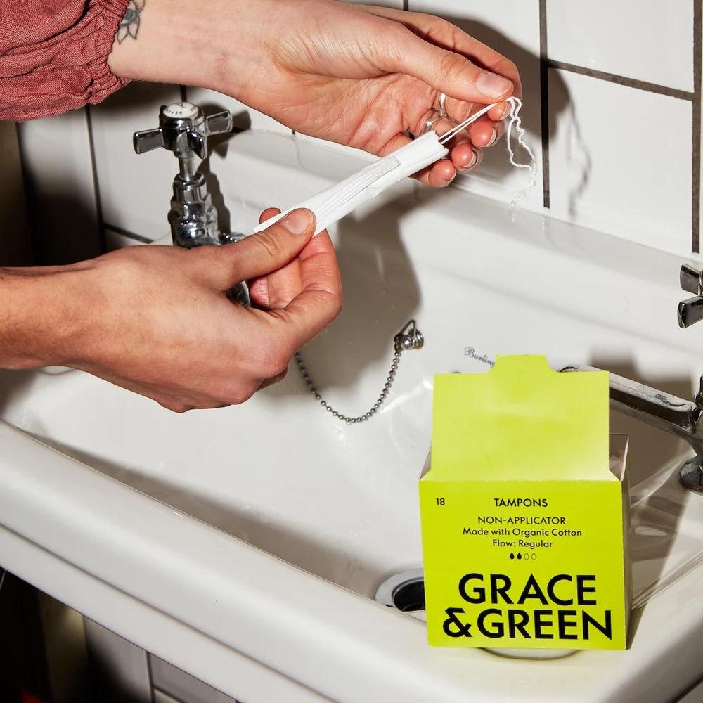 Tampoane bumbac organic alb, detaliu textură, cutie verde deschis, chiuvetă albă apropiere.