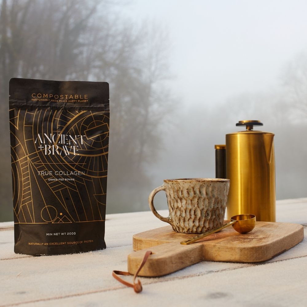 Pungă colagen Ancient and Brave pe masă rustică, alături de cană texturată și carafă aurie.