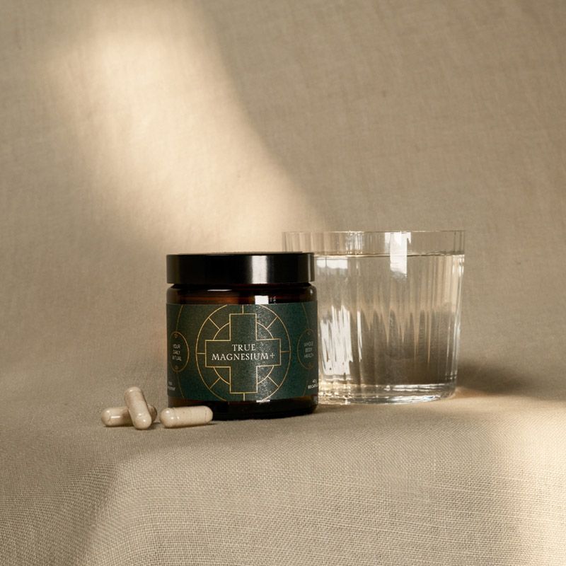 Borcan brun de True Magnesium+ cu etichetă verde, capsule albe și pahar cu apă pe material texturat bej.