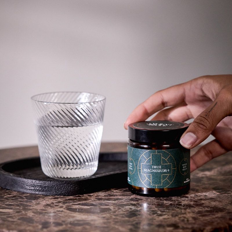 Borcan True Magnesium+ cu etichetă verde, lângă pahar cu apă pe tăviță neagră, pe masă de marmură.