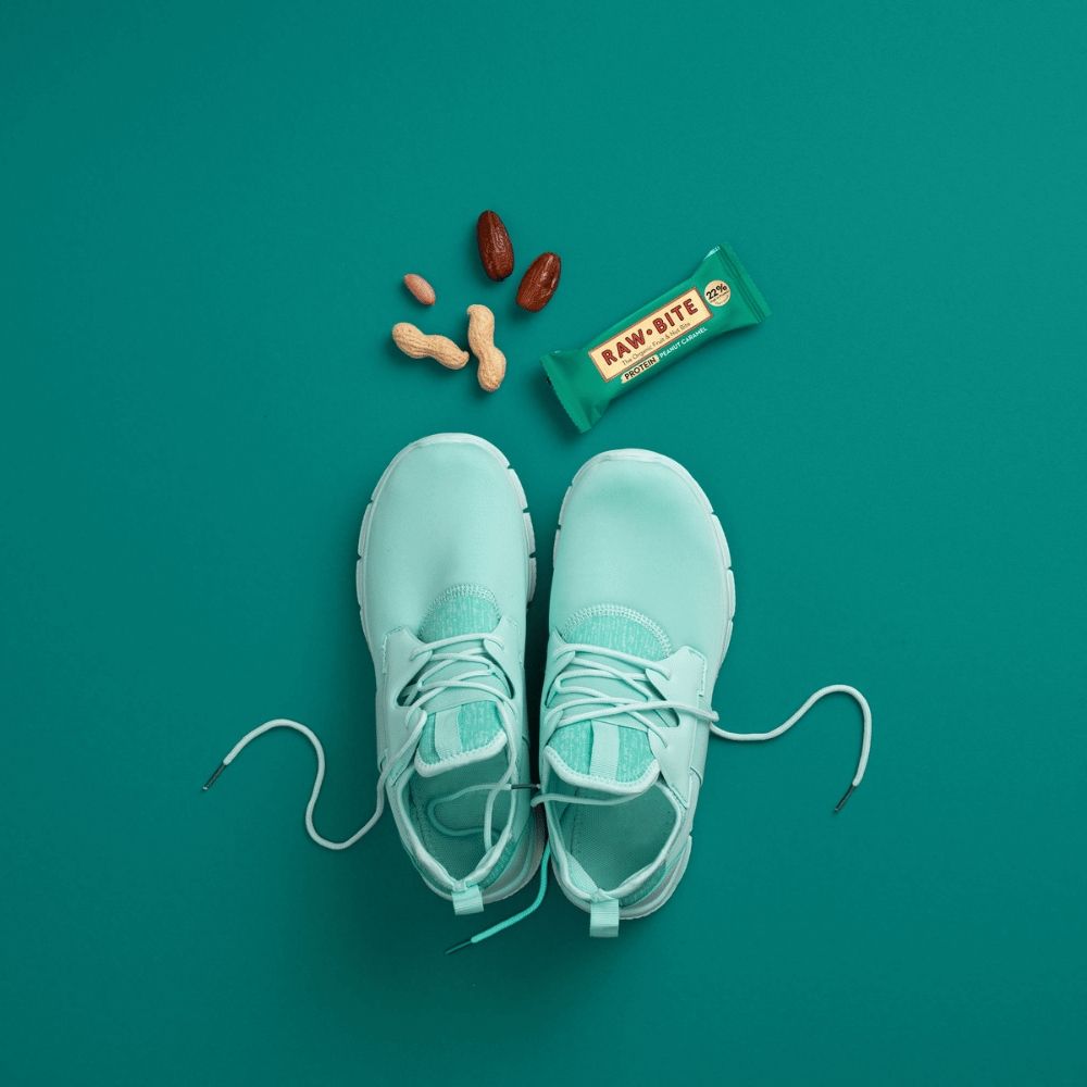 Baton proteic Raw Bite peanut caramel, verde, 45g, alături de alune și curmale pe fundal turcoaz.