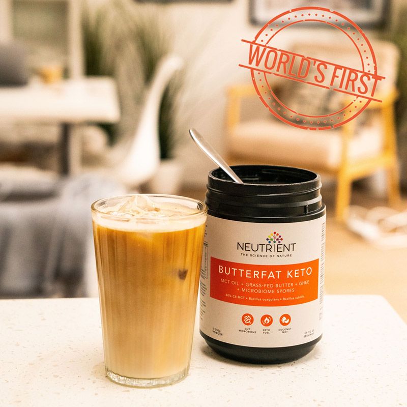 Pudră Butterfat Keto într-un borcan negru, linguriță și pahar de cafea cu gheață pe masă.