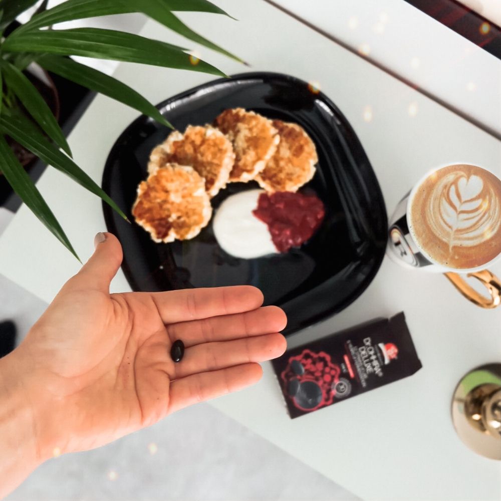 Capsula neagră Dr.Ohhira® pe mână, lângă preparat pe farfurie neagră și cafea cu model latte art.