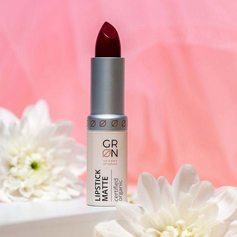 Ruj mat roșu Bio Organic, tub argintiu și bej, fundal roz, flori albe decor.