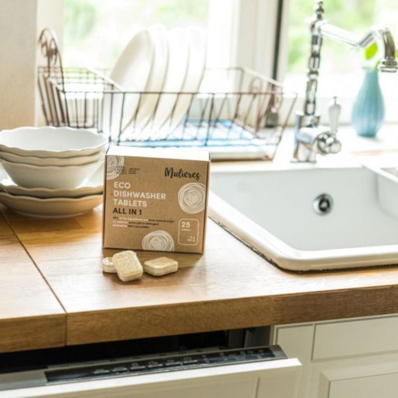 Tablete ecologice Mulieres pe blat de lemn, cutie carton maro, chiuvetă albă, veselă albă în fundal.