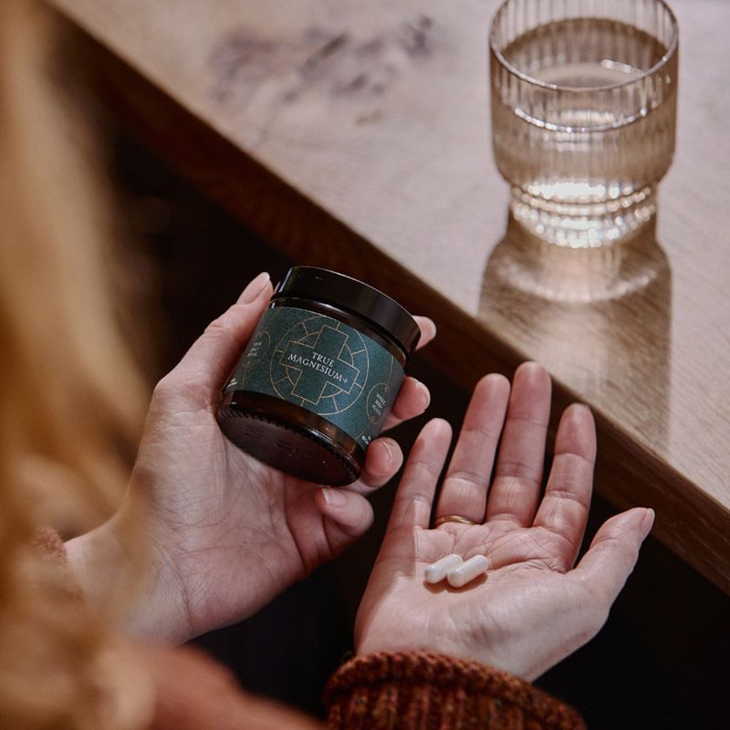 Supliment capsule True Magnesium+ în mână, ambalaj verde închis, capsule albe, detalii etichetă vizibile.
